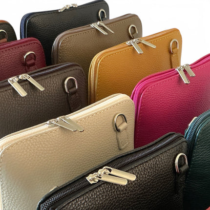 A close-up, overlapping product shot of multiple pebbled leather zip-around crossbody bags in different colors, including black, dark brown, dark tan, wine, cream and teal, arranged on a white background.