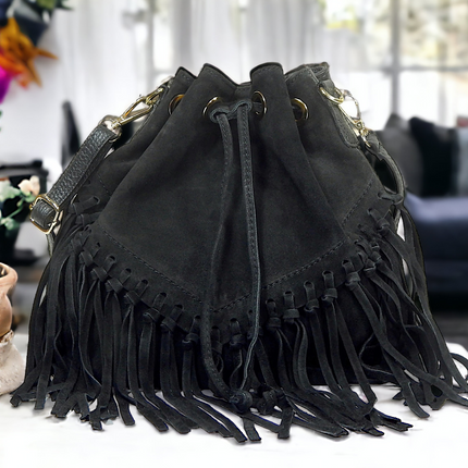 Front view of black leather and suede bucket bag with fringes on a table with plant. Drawstring closure. Room with furniture in the background