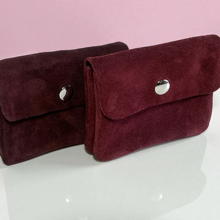 Two burgundy suede card holders and coin purses with silver buttons on a white surface and pink background